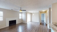 This single-floor space is designed with a neutral, contemporary aesthetic throughout. The spacious living room serves as the centerpiece, showcasing rich hardwood-style flooring, a textured ceiling, and a ceiling fan with integrated lighting. A classic fireplace with a white tile surround adds a focal point to the room, while vertical blinds over a sliding door and an additional window allow for natural light. The open layout connects the living area to the kitchen, which features white cabinetry, granite-look countertops, a peninsula with an undermount sink, stainless steel appliances including an electric range with a built-in range hood, and a geometric patterned tile backsplash. The bedroom offers hardwood-style flooring, a textured ceiling with a vent, and a window with blinds providing natural light. A hallway leads to the bathroom, which is finished with a vessel sink atop a white vanity with granite countertop, a framed mirror, and hardwood-style flooring. A closet with bifold doors is accessible nearby. The unit maintains a cohesive, neutral color palette with warm flooring tones throughout.