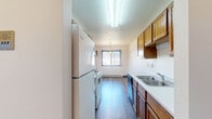 Located on Ninth Avenue Circle South in Fargo, North Dakota, this two-bedroom, one-bathroom apartment offers a functional and practical single-floor layout. The galley-style kitchen is well-equipped with warm oak cabinetry, a double sink, dishwasher, electric range, and a full-size refrigerator, all set above durable hardwood-style flooring. The kitchen flows directly into a combined dining and living area, where a pendant light fixture and a window with natural light create a welcoming atmosphere. Baseboard heating is found throughout the unit, ensuring comfort during North Dakota winters, and a wall-mounted air conditioning unit provides relief in warmer months. Both bedrooms feature plush carpeting, large windows dressed with vertical blinds, and ample closet space with bi-fold doors — the primary bedroom being notably spacious. The full bathroom includes a tub-shower combination with clean white surroundings. Additional storage is provided by four closets spread throughout the unit, along with a dedicated utility room. Textured ceilings are present throughout, and warm wood-toned trim and door frames lend a cohesive, classic character to the space. With a straightforward, efficient layout, this apartment offers comfortable, practical living in a residential Fargo neighborhood.