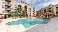 This property combines well-appointed interior living spaces with exceptional outdoor amenities. The centerpiece of the community is a generously sized resort-style pool, framed by a wide surrounding deck furnished with lounge chairs and shaded by umbrellas. Towering palm trees and lush landscaping enhance the outdoor ambiance, while the warm-toned Mediterranean-inspired building exterior provides an elegant backdrop.

Inside, the open-plan kitchen and living area showcases rich hardwood-style flooring, crown molding, and abundant natural light streaming through large windows dressed with teal curtains. The kitchen is outfitted with dark espresso cabinetry, granite countertops, a kitchen island, and a full suite of stainless steel appliances, including a refrigerator with ice maker, dishwasher, microwave, and range. Track lighting and a ceiling fan add both function and style to the connected living space.

The entryway features hardwood-style flooring, a security keypad, and direct access to the kitchen. Neutral-toned walls throughout are accented with framed artwork, contributing to a polished, cohesive aesthetic. The overall atmosphere is modern, comfortable, and resort-inspired.