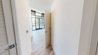 This contemporary single-floor unit is defined by its striking architectural design, most notably the expansive floor-to-ceiling black-framed windows that flood the main living area with natural light and frame open outdoor views of surrounding greenery. The living space is open and airy, with light-toned hardwood flooring that flows seamlessly throughout the unit, enhancing its clean, modern aesthetic. Large sliding glass doors provide direct access to a private balcony, extending the living area outdoors.

The kitchen is thoughtfully designed with high-gloss white cabinetry reaching to the ceiling, white quartz countertops, a stainless steel sink, and a full suite of stainless steel appliances including a range, over-the-range microwave, dishwasher, and refrigerator. A kitchen island adds additional counter and prep space.

The bathroom features a clean, minimalist design with a single-sink vanity topped in white, a toilet, and a combined shower and soaking tub with neutral-toned tile surround. A grab bar adds a functional safety element.

Storage is well-addressed through a built-in closet with a sliding panel door, offering dual hanging rods and a full shelving system. Throughout the unit, crisp white walls and recessed lighting contribute to a bright, polished, and cohesive modern atmosphere.