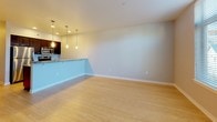 This single-floor space offers a clean, contemporary design with a cohesive aesthetic carried throughout. Light hardwood flooring runs across the main living and sleeping areas, lending warmth and visual continuity to the open layout. The living area is generously proportioned, with neutral-toned walls and a bright, airy feel that creates a versatile canvas for furnishing.

The galley-style kitchen is well-appointed, featuring dark espresso cabinetry paired with speckled granite countertops that extend to a peninsula counter overlooking the living area. Stainless steel appliances, including a refrigerator, range, and built-in microwave, are integrated seamlessly into the cabinetry. Pendant lighting fixtures hang above both the peninsula and the main kitchen workspace, providing focused illumination while adding a modern design touch. Recessed ceiling lights further enhance the kitchen's functionality.

The bathroom continues the granite countertop theme, with a dark vanity cabinet, an undermount sink, and a large wall mirror. A shower/tub combination and neutral tile work complete the space. A dedicated bedroom area with a closet alcove provides practical storage. Overall, the unit maintains a consistent, modern palette throughout.