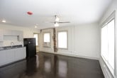 This single-floor unit presents a compact yet functional layout with a series of clearly defined living spaces. The main living area is highlighted by dark-stained hardwood floors that run throughout, a ceiling fan overhead, and windows fitted with horizontal blinds that allow natural light to filter in. An exposed brick section along one wall adds a raw, urban character to the space. An in-unit washing machine is conveniently positioned within the main room, providing practical laundry access. The kitchen is efficiently arranged with white cabinetry, dark countertops, and recessed lighting. It is equipped with a full suite of black appliances, including a gas range, over-the-range microwave, and a full-size refrigerator. A single window in the kitchen area brings in additional natural light. The bathroom is cleanly appointed with a walk-in shower stall, a wall-mounted sink, a mirror, and a toilet — all finished in neutral tones for a simple, functional aesthetic. The overall layout of the unit, as seen in the dollhouse view, is open and straightforward, making efficient use of the available floor space across a single level.