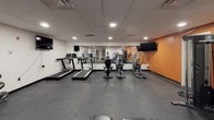 This single-floor fitness center is designed to support a well-rounded workout experience. The space is finished with durable speckled rubber flooring throughout, providing both safety and functionality for a range of exercise activities. A drop ceiling with recessed lighting ensures consistent, bright illumination across the entire area.

The cardio zone features multiple treadmills and elliptical machines lined along a mirrored wall, which visually expands the space and allows users to monitor their form during exercise. Wall-mounted flat-screen televisions are positioned at multiple points around the room, providing entertainment during workouts.

The strength training area includes an Inspire cable machine system, an adjustable workout bench, a dumbbell rack, and a medicine ball stand, offering a variety of resistance and functional training options. A designated open floor area provides room for stretching or bodyweight exercises.

Bold orange-painted concrete block walls serve as a defining design element, lending the space an energetic and motivating atmosphere. Glass-paneled doors and interior windows connect the fitness center to an adjacent area, adding a sense of openness. The layout is practical and efficient, making effective use of the available space for a comprehensive fitness facility.
