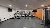 This single-floor fitness center is designed to support a well-rounded workout experience. The space is finished with durable speckled rubber flooring throughout, providing both safety and functionality for a range of exercise activities. A drop ceiling with recessed lighting ensures consistent, bright illumination across the entire area.

The cardio zone features multiple treadmills and elliptical machines lined along a mirrored wall, which visually expands the space and allows users to monitor their form during exercise. Wall-mounted flat-screen televisions are positioned at multiple points around the room, providing entertainment during workouts.

The strength training area includes an Inspire cable machine system, an adjustable workout bench, a dumbbell rack, and a medicine ball stand, offering a variety of resistance and functional training options. A designated open floor area provides room for stretching or bodyweight exercises.

Bold orange-painted concrete block walls serve as a defining design element, lending the space an energetic and motivating atmosphere. Glass-paneled doors and interior windows connect the fitness center to an adjacent area, adding a sense of openness. The layout is practical and efficient, making effective use of the available space for a comprehensive fitness facility.