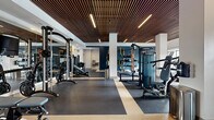 Located at 400 Buford Hwy in Suwanee, GA, this well-appointed fitness center is thoughtfully divided into two dedicated exercise rooms, offering a comprehensive workout experience across a generously sized single-floor layout. A distinctive slatted wood ceiling runs throughout the space, adding warmth and architectural character to the otherwise crisp, modern interior defined by white walls and neutral flooring with geometric color-block detailing.

The main exercise room is a strength and cardio hub, equipped with cable machines, weight benches, free weights, and a punching bag for functional training. Full-length mirrors line the walls, amplifying the sense of space while allowing users to monitor their form. A row of treadmills and elliptical machines lines a wall of large windows, inviting natural light and outdoor views to energize cardio sessions.

The second exercise room provides a dedicated space for group-style and low-impact training, furnished with spin bikes, rowing machines, BOSU balls, stability balls, and dumbbells. A floor-to-ceiling mirror and large windows overlooking what appears to be a pool area enhance the bright, open atmosphere.

Accessory areas feature organized racks stocked with medicine balls, kettlebells, and exercise mats, ensuring all equipment is readily accessible. The facility presents a clean, professional ambiance suited to a wide range of fitness goals.
