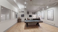 Located at 250 W 5th St in downtown Cincinnati, this single-floor lounge space blends industrial architecture with contemporary design to create a versatile social environment. The room is defined by its soaring ceilings, exposed piping, and track lighting suspended overhead, lending it a loft-like, urban character. Light hardwood flooring runs throughout, contributing warmth and continuity to the space.

At the center of the room sits a substantial pool table, serving as the focal point of the entertainment area. Surrounding it is ample open floor space, allowing for easy movement and socialization. A dedicated lounge area features a plush gray sectional sofa, an upholstered leather accent chair, a round coffee table, and a wall-mounted flat-screen TV, creating a comfortable retreat for relaxing or watching content.

A built-in wet bar along one wall showcases dark cabinetry, a light countertop, a stainless steel mini-refrigerator, and a decorative patterned wallcovering backdrop, making it well-suited for entertaining. Wall-mounted sconces flank the television above the bar, adding ambient warmth. Industrial-style framed window panels along the walls enhance the architectural character, while frosted-glass interior doors provide access points throughout. The overall ambiance is modern, refined, and well-appointed.