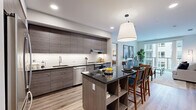 This well-appointed 2-bedroom, 2-bathroom residence offers a cohesive modern aesthetic across a single floor. The entrance leads into an open-concept living and kitchen area bathed in natural light. The kitchen is a standout feature, equipped with sleek, textured dark cabinetry, stainless steel appliances including a refrigerator, dishwasher, and range with hood, complemented by a white subway tile backsplash and a central island with a dark granite countertop and built-in shelving — ideal for casual dining. The living room flows seamlessly from the kitchen and opens to a private balcony through large sliding glass doors, offering views of the surrounding community. The primary bedroom is generously sized, featuring large windows with exterior views and direct access to a well-finished en-suite bathroom with a double-sink vanity, dark cabinetry, an illuminated mirror, and a separate shower area. The second bedroom is equally bright with ample window space. Both bathrooms are finished with wood-look luxury vinyl plank flooring consistent throughout the residence. Two closets provide excellent storage, and the layout is completed by a welcoming entrance area. The overall ambiance is clean, contemporary, and functional.