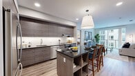 This well-appointed 2-bedroom, 2-bathroom residence offers a cohesive modern aesthetic across a single floor. The entrance leads into an open-concept living and kitchen area bathed in natural light. The kitchen is a standout feature, equipped with sleek, textured dark cabinetry, stainless steel appliances including a refrigerator, dishwasher, and range with hood, complemented by a white subway tile backsplash and a central island with a dark granite countertop and built-in shelving — ideal for casual dining. The living room flows seamlessly from the kitchen and opens to a private balcony through large sliding glass doors, offering views of the surrounding community. The primary bedroom is generously sized, featuring large windows with exterior views and direct access to a well-finished en-suite bathroom with a double-sink vanity, dark cabinetry, an illuminated mirror, and a separate shower area. The second bedroom is equally bright with ample window space. Both bathrooms are finished with wood-look luxury vinyl plank flooring consistent throughout the residence. Two closets provide excellent storage, and the layout is completed by a welcoming entrance area. The overall ambiance is clean, contemporary, and functional.