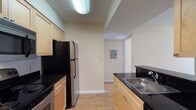 This single-floor space features a galley-style kitchen as a central highlight, designed with practicality in mind. The kitchen is outfitted with light wood cabinetry that provides ample storage on both sides of the corridor-style layout. Dark granite-look countertops contrast the cabinetry, lending a polished appearance, while the stainless steel sink and black appliances — including a refrigerator and electric range — complete the functional setup. Overhead, a flush fluorescent light fixture ensures the kitchen is well-lit, complementing the textured ceiling finish found throughout the space. The flooring features light wood-tone laminate or vinyl planking, adding warmth while remaining easy to maintain. Neutral beige walls keep the atmosphere clean and versatile. An electrical panel is visible along the hallway wall beyond the kitchen, indicating practical utility access. The hallway connects the kitchen to additional areas of the unit, with a doorway visible at the far end suggesting further living or storage space. The overall design is straightforward and functional, with a cohesive, neutral aesthetic throughout.