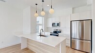 This thoughtfully designed single-floor studio offers a clean, contemporary aesthetic throughout. The open-concept layout seamlessly connects the kitchen and living area, maximizing functionality within the compact footprint. The kitchen serves as a standout centerpiece, featuring a large island with a white quartz countertop and a light wood-paneled base, an undermount sink with a matte black faucet, and a full suite of stainless steel appliances including a French door refrigerator, electric range, built-in microwave, and dishwasher. White shaker-style cabinetry extends to the ceiling, providing generous storage. Three globe pendant lights with black and gold accents hang above the island, adding a stylish focal point. Light-toned hardwood flooring runs throughout, enhancing the airy, bright atmosphere. The living area is bathed in natural light from expansive floor-to-ceiling windows and a sliding glass door that opens to a private balcony, offering sweeping urban and mountain views. A closet provides dedicated storage. The space maintains a crisp, neutral palette of whites and soft naturals, creating an inviting and versatile environment ideal for modern living.
