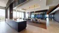 This refined, single-floor space exemplifies contemporary commercial design, seamlessly blending functionality with upscale aesthetics. The open-plan layout is anchored by large-format tile flooring that transitions gracefully into rich hardwood, creating distinct zones throughout. Floor-to-ceiling glass walls flood the interior with natural light while framing expansive panoramic city views from multiple vantage points.

The main gathering area features a sleek dark-cabinet island with a pristine countertop, complemented by sculptural branch-style pendant chandeliers and spherical globe fixtures that add dramatic visual interest. A plush lounge zone with neutral upholstered seating and a wall-mounted display provides a comfortable setting for relaxed collaboration.

Multiple glass-enclosed conference rooms are thoughtfully positioned throughout the space, each outfitted with ergonomic seating, integrated display screens, and statement lighting fixtures. A smaller private meeting room showcases bold abstract artwork and frosted glass partitions for added privacy.

The kitchen area is generously sized, centered around a large island with bar seating, a built-in gas cooktop, and stainless steel appliances. Curated artwork and sculptural décor pieces, including a striking organic wood sculpture, are displayed throughout the corridors, lending a gallery-like quality to the overall ambiance.