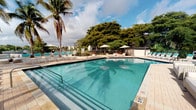 This property's most prominent feature is its generously sized outdoor swimming pool, finished in vivid turquoise water that reflects the surrounding palm trees and open sky. The pool deck is outfitted with a collection of lounge chairs and shade umbrellas, creating a comfortable environment for relaxation. Mature tropical trees and swaying palms frame the space, lending a lush, resort-like ambiance to the outdoor area. A safety fence borders the perimeter, separating the pool area from what appears to be a waterway beyond, offering scenic views of the natural surroundings. A small poolside structure, likely a restroom or storage facility, is visible at the rear of the deck, adding functional convenience to the space. The overall setting evokes a warm, sun-drenched atmosphere characteristic of South Florida. The single-story layout of the property contributes to an accessible, open feel throughout. Unfortunately, the interior images provided are too underexposed to discern specific details about the indoor spaces, so the description is limited to the clearly visible outdoor amenities that define this property's character and appeal.