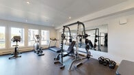 Located at 487 Broad St in Halifax, Nova Scotia, this fitness room offers a compact yet comprehensively equipped exercise space across a single floor. The room features crisp white walls, recessed lighting, and durable rubber flooring throughout, creating a clean and functional environment suited for a variety of workout routines.

Natural light fills the space through large windows along one wall, providing views of the surrounding neighbourhood and an open, airy atmosphere. A full-wall mirror on one side visually expands the room while allowing users to monitor their form during strength training.

The cardio section is well-appointed with a True treadmill, an elliptical trainer, and a rowing machine, all positioned to take advantage of the natural light from the windows. On the strength training side, the room houses a Smith machine with an adjustable bench, a Hoist functional cable trainer, a rack of dumbbells in varying weights, kettlebells, and additional free weight plates. A stability ball and balance disc round out the functional training options.

Climate comfort is maintained via a wall-mounted mini-split air conditioning unit, ensuring a comfortable workout environment year-round. The space is efficiently laid out to maximize usability while maintaining clear movement pathways, making it a practical and well-organized fitness facility.