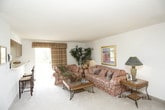 This single-floor residence presents a cohesive and comfortable living environment across several well-defined spaces. The living room serves as the central gathering area, featuring plush floral upholstered sofas, warm ambient lighting from stylish table lamps, and decorative indoor plants that add a natural touch. Framed artwork adorns the walls, contributing to an inviting atmosphere with neutral carpet flooring throughout.

The dining area is equipped with a glass-top table surrounded by woven chairs, accented by a ceiling fan with integrated lighting overhead. Adjacent to it, the kitchen provides functional cabinetry in warm wood tones, ample counter space, and a coil-burner electric range with an overhead ventilation hood.

The bedroom is generously sized, featuring a decorative wrought iron bed frame, coordinated wood dresser with mirror, and multiple windows dressed with patterned curtains that allow natural light to filter in. A spacious open closet offers practical storage, and framed artwork decorates the neutral walls.

The bathroom includes a full tub and shower combination with sliding frosted glass doors, a toilet, and vanity, all finished in clean, light tones that contribute to the space's overall bright and tidy presentation.