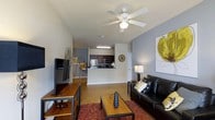 This single-floor residence offers a thoughtfully designed open-concept layout that brings together the living area and kitchen in a cohesive, functional arrangement. The living room serves as the heart of the space, featuring warm hardwood-style flooring, a ceiling fan with integrated lighting, and neutral-toned walls accented with tasteful artwork. A comfortable seating area centers around a coffee table and is complemented by a media console, creating an inviting atmosphere for both relaxing and entertaining. The kitchen is positioned at the rear of the open floor plan and features dark cabinetry paired with stainless steel appliances, including a refrigerator and microwave. A raised breakfast bar provides a convenient transition between the kitchen and living space, ideal for casual dining or entertaining guests. The layout maximizes the sense of openness, with clean sightlines extending from the front of the living area through to the kitchen. The overall ambiance is modern and casual, with a neutral color palette punctuated by warm accent tones. The space is well-lit with a combination of natural and recessed lighting, contributing to a bright and welcoming environment throughout.