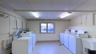 This single-floor laundry facility is designed for practical, high-capacity use. The room is equipped with multiple washing machines and dryers arranged along both side walls, providing ample capacity for shared or communal use. The layout is open and efficiently organized, allowing easy access to all appliances.

The space features a drop ceiling fitted with fluorescent lighting fixtures that illuminate the room evenly, creating a bright and functional environment. The walls are finished in a clean, neutral tone, and the flooring is a light-colored, easy-to-maintain surface suited to the demands of a laundry space.

A centrally positioned window on the far wall allows natural light to enter and provides a view of the exterior parking area, adding a degree of openness to the otherwise utilitarian space. Visible utility infrastructure, including ventilation ductwork running along the ceiling, reflects the room's functional design.

Overall, the space is straightforward and purposefully arranged, prioritizing accessibility and efficiency for laundry operations.