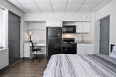 This 1-bedroom, 1-bathroom unit offers a practical and efficient layout across a single floor. The open-concept living and dining area flows seamlessly into the kitchen, creating a cohesive and functional common space. The living area features wood-look flooring, a drop tile ceiling, and floor-to-ceiling windows that allow for generous natural light and urban outdoor views. A wall-mounted air conditioning unit provides climate control directly in the living space.

The kitchen is equipped with grey cabinetry on both upper and lower sections, a double-basin stainless steel sink, laminate countertops, and a white dishwasher. A curved breakfast bar counter separates the kitchen from the living and dining area, adding a convenient casual dining option.

The bedroom is a well-proportioned space featuring the same wood-look flooring and drop tile ceiling, with expansive windows on two walls that flood the room with natural light and provide open exterior views. A built-in closet offers dedicated storage.

The bathroom includes a vanity with a speckled countertop, an integrated sink, a large mirror, and an open closet alcove with a shelf and hanging rod for additional storage. The unit's consistent design elements, including neutral tones, wood-look floors, and grey accents, give it a cohesive and clean aesthetic throughout.
