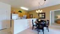 This single-floor space showcases an open-concept layout that seamlessly connects the kitchen and dining areas, creating a functional and inviting environment. The kitchen features light wood cabinetry accented by warm yellow walls, offering generous storage with upper and lower cabinets. White appliances, including a refrigerator and microwave, complement the bright, clean aesthetic. A white half-wall partition elegantly separates the kitchen from the adjoining dining area while maintaining an open sightline throughout the space. The dining area is anchored by a round dark wood table surrounded by matching black X-back chairs, lending a classic, grounded feel to the room. Above, a wrought iron chandelier with fabric shades provides warm, focused lighting. A framed mirror and wall art add decorative character to the dining space. The neutral carpeted flooring flows throughout, contributing to the overall warmth of the interior. The consistent use of warm tones, from the golden yellow walls to the wood finishes, creates a cohesive and welcoming ambiance throughout this efficiently designed single-floor space.