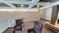 This single-floor commercial space makes a memorable first impression through its carefully curated design elements and refined architectural details. At the heart of the lobby stands a dramatic centerpiece installation featuring natural white birch trees arranged between dark wood-clad columns, set atop a stone base with decorative river rocks and illuminated to create a warm, organic focal point. Paired leather bench seating flanks the installation, inviting visitors to pause and take in the surroundings.

The architectural ceiling treatment is a standout feature, composed of a geometric coffered design with wood-paneled recessed sections and integrated recessed lighting that lends depth and sophistication to the overall aesthetic. Warm ambient LED strip lighting runs along the upper walls, enhancing the sense of height and refinement.

A seating lounge area occupies one portion of the space, furnished with plush upholstered armchairs in a muted gray-purple tone and accented with glass side tables. The walls in this zone are clad in large-format stone block tiles in a neutral cream palette, adding texture and a grounded, substantial character. Soft, patterned carpet underfoot ties the space together with warmth. Access to additional office or meeting rooms is visible through glass-paneled doors, suggesting a functional, professional environment beyond the welcoming lobby.