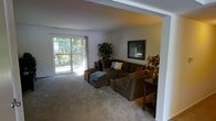 This single-floor residence offers a welcoming and well-appointed living space designed for comfort and functionality. The living room serves as the heart of the home, featuring soft, neutral-toned carpeting that extends throughout, creating a warm and cohesive atmosphere. A large sliding glass door floods the room with natural light and provides direct access to a private balcony, where mature trees and greenery create a serene, nature-filled backdrop. Vertical blinds frame the door, offering flexible light control throughout the day. The living area is generously proportioned, easily accommodating a substantial sectional sofa and additional seating, making it ideal for both relaxing and entertaining. Structural beams accent the ceiling, adding architectural character to the space. Decorative elements, including framed wall art and indoor plants, contribute to a lived-in, inviting ambiance. A textured ceiling adds subtle visual interest overhead. The open layout connects seamlessly to adjacent areas of the home, enhancing the overall sense of flow. The space strikes a balance between practical design and comfortable living, with natural light and outdoor connectivity as standout features.