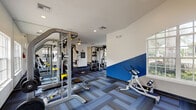 This fitness center is a thoughtfully designed, single-level facility offering a comprehensive workout environment. The space is divided into distinct functional zones, each outfitted with dedicated equipment. The cardio area houses multiple treadmills, elliptical trainers, and a Concept2 rowing machine, all positioned near large windows that frame views of the surrounding greenery and pool area, filling the space with natural light.

The strength training zone features a Paramount cable machine system, a Hoist multi-station unit, free weights, and various resistance machines, providing options for full-body conditioning. A dedicated stretching and flexibility area includes ab equipment and floor space for functional training.

Architecturally, the facility stands out with vaulted ceilings accented by arched transom windows and ceiling fans, creating an open, airy atmosphere. Bold royal blue geometric wall accents add a dynamic visual element throughout. Patterned modular carpet tiles in blue and grey tones cover the floors, providing both comfort and style.

A kitchenette area with white cabinetry and a sink offers added convenience, while louvered storage doors and recessed lighting contribute to the clean, functional design. French doors in one section open directly to the outdoors, enhancing the connection between the interior workout space and the exterior environment.