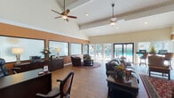 Located at 25 Pelham Rd in Greenville, SC, this single-story community clubhouse offers a warm and inviting atmosphere designed for both relaxation and socializing. The main lounge is the heart of the space, distinguished by dramatic vaulted ceilings with tongue-and-groove detailing and dual ceiling fans that enhance airflow throughout. Rich hardwood flooring, warm terracotta accent walls, and an abundance of natural light from floor-to-ceiling glass doors and windows create a welcoming ambiance. Comfortable seating arrangements — including leather sofas, upholstered armchairs, and a circular seating area anchored by a patterned area rug — offer multiple zones for gathering. A flat-screen TV and framed artwork add to the lived-in, curated feel of the lounge. Large glass doors open directly to an outdoor pool and patio area, seamlessly blending indoor and outdoor living. A dedicated café bar area, styled as "Café Bar Bonito," features rich cherry cabinetry, a full countertop with a double sink, a dishwasher, a coffee station, and a water cooler, making it ideal for resident events and daily refreshments. A smaller lounge nook with leather barrel chairs and a decorative mirror provides a quieter retreat within the space.