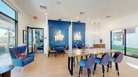 Located at 6755 W McDowell Rd in Phoenix, Arizona, this single-floor amenity space combines a fully equipped exercise room with a vibrant dining and lounge area, offering a well-rounded community environment. The fitness center features a bold blue-and-white geometric accent wall, carpet tile flooring, and an impressive array of equipment including treadmills, cable machines, a punching bag, free weights, and cardio machines. Large floor-to-ceiling windows and glass garage-style doors flood the gym with natural light while providing outdoor views of the surrounding grounds. Ceiling fans and linear pendant lighting enhance comfort throughout. The adjoining dining area is designed with equal care, showcasing light hardwood-style flooring, crisp white walls, and striking cobalt blue accent walls that define cozy nook seating with individual tables, caramel leather chairs, and pendant lights overhead. A large communal table with a colorful striped top and modern blue upholstered chairs serves as a central gathering point. Expansive glass windows and sliding doors open the dining area to outdoor views of palm trees and green space, bathing the room in natural light. A wet bar area with cabinetry adds functional convenience. The space is cohesive in its modern, energetic aesthetic throughout.
