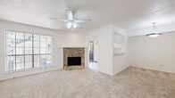 This two-story residence offers a thoughtfully organized layout spread across two floors. The main level features a bright living room anchored by a striking stone fireplace with a chevron-detailed wood surround, complemented by a ceiling fan, textured ceiling, and large windows that allow natural light to fill the carpeted space. Adjacent to the living room, the kitchen is finished with white cabinetry, tile flooring, a full suite of white appliances including a refrigerator, range, and built-in microwave, along with a sink and ample counter space. A pass-through opening connects the kitchen to the living area, enhancing the open feel of the main floor. Two bathrooms serve the home, one of which features a single-sink vanity with white cabinetry, a large mirror, tile flooring, and a tub/shower combination. The upper floor houses the bedroom, a generously sized and carpeted retreat with a ceiling fan, vertical blind-covered window, and direct access to a walk-in closet with built-in shelving. Throughout the home, neutral tones, carpet in living and sleeping areas, and tile in wet zones create a clean, cohesive aesthetic. Staircase access connects both levels seamlessly.