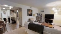 This single-floor residence offers a comfortable and thoughtfully arranged living environment. The living area serves as the heart of the home, featuring neutral white walls that create a clean, bright backdrop for the space. Plush seating, including a light-toned sofa accented with a dark throw blanket, anchors the room and invites relaxation. A well-stocked wooden bookshelf filled with books and personal mementos adds character and warmth, while a flat-screen television on a matching media stand provides a functional entertainment setup. A striking piece of wall art depicting a grand piano adds a bold, artistic focal point to the room, complementing the overall curated aesthetic. A floor lamp provides warm ambient lighting, enhancing the inviting atmosphere. The flooring transitions from hardwood in the connecting hallway to soft carpet in the main living area, adding texture and comfort underfoot. An open doorway leads to what appears to be a bedroom, offering a glimpse into the private quarters beyond. The layout feels connected and accessible, with the single-floor design ensuring ease of movement throughout. Overall, the space strikes a balance between functional living and personal expression, making it a warm and welcoming home environment.