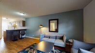 This single-floor residence offers a thoughtfully arranged layout with two bedrooms, a full bathroom, a galley-style kitchen, and a welcoming living area. The living room makes a strong first impression with its bold slate-blue accent wall, warm wood-look flooring, and a cozy seating arrangement anchored by a vibrant geometric-patterned area rug. A framed wall mirror and a sleek arc floor lamp add character and depth to the space, while a window allows natural light to filter in near the entry door. The galley kitchen is efficiently designed with dark wood cabinetry lining both sides, light countertops, a white electric range, and a full-size refrigerator, complemented by a window that brings in additional natural light. The two bedrooms each feature the same warm wood-look flooring and textured ceilings. One bedroom includes a built-in desk and shelving unit, offering a practical workspace, while the other features a walk-in closet, a ceiling fan, and windows with blinds for light control. The bathroom is outfitted with a full tub-shower combination with tiled surround, a toilet, and a vanity with a countertop sink. An in-unit washer is also visible, adding everyday convenience to this functional and comfortable space.