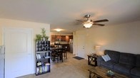 This single-floor residence offers a thoughtfully designed open-concept layout that seamlessly connects the main living areas. The living room provides a comfortable, welcoming atmosphere, complete with a ceiling fan that adds both function and a classic touch to the space. Warm neutral-toned walls and plush carpeting throughout contribute to an inviting ambiance.

The living area flows naturally into a defined dining space, which accommodates a dining table and chairs, making it ideal for everyday meals or entertaining guests. A bookshelf and wine rack unit serves as a subtle, decorative divider between the living and dining zones, adding character and practical storage without fully closing off the open feel.

The kitchen is located at the rear of the main living area and features rich, dark wood cabinetry paired with stainless steel appliances, including a refrigerator, stove, and microwave. Tile flooring in the kitchen provides a clean, durable surface that contrasts nicely with the carpeted living areas. A door near the kitchen leads to an additional room or entry point, adding to the unit's functional flow. Overall, the space balances practicality with a cozy, lived-in warmth throughout its single level.