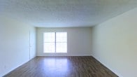 This single-floor space presents a clean, open layout with a straightforward and functional design. The main living area showcases dark hardwood-style flooring that contrasts attractively with the light-colored walls, creating a balanced and inviting atmosphere. A centrally positioned window allows generous amounts of natural light to filter into the room, brightening the interior and lending an airy quality to the space. The textured ceiling adds a subtle decorative element overhead, contributing modest visual interest to the otherwise minimalist aesthetic. Neutral wall tones provide a versatile backdrop, well-suited to a variety of décor styles. A white door is visible along one wall, offering access to additional areas of the unit. The baseboards throughout are clean and well-maintained, complementing the overall simplicity of the design. The open floor plan allows for flexible furniture arrangement, making it adaptable for various uses. Overall, the space offers a practical and understated environment, highlighted by natural light and durable flooring finishes.