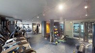 This fitness center offers a comprehensive workout environment designed to accommodate a wide range of exercise routines. The space is laid out across a single floor with distinct zones for various training needs. A dedicated cardio area lines one side of the room, featuring multiple treadmills and other aerobic machines, all positioned to make efficient use of the available floor space. Strength training equipment and cable machines occupy a central zone, providing options for full-body resistance workouts. A free weights section includes a well-organized dumbbell rack stocked with a colorful variety of medicine balls and hand weights, accompanied by benches for seated or supported exercises. Large floor-to-ceiling mirrors are strategically placed to allow users to monitor their form throughout their workouts. A frosted glass partition with a decorative motif adds a design element while subtly defining separate areas within the space. Track lighting runs along the ceiling, providing bright, even illumination throughout. The flooring consists of durable dark tiles suited for high-traffic fitness use. The overall ambiance is modern and functional, with clean lines and a neutral color palette accented by warm tones on select walls, creating an energizing yet focused atmosphere.
