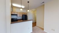 This single-floor residence is thoughtfully laid out to maximize functionality and flow. The kitchen serves as a central hub, featuring warm-toned wood cabinetry, dark laminate countertops, and black appliances including an electric range and refrigerator. A double stainless steel sink sits along one wall, while a breakfast bar peninsula separates the kitchen from the adjacent living and dining areas. Pendant lights hang above the bar and dining space, where a chandelier adds a finishing touch overhead.

The open layout connects the kitchen to the carpeted living and dining areas, creating a cohesive and airy feel throughout. Soft neutral tones on the walls complement the light carpet found in the living spaces and bedrooms. The bedrooms are generously proportioned, each featuring windows with blinds that allow natural light to filter in. One bedroom includes a built-in bench beneath its window, adding a unique architectural detail. Recessed lighting and standard ceiling fixtures provide consistent illumination throughout.

The bathroom is finished with tile flooring, a wood-fronted vanity with a light countertop, and a full-size soaking tub with a shower. Hallways connect the various rooms efficiently, with multiple paneled interior doors maintaining a consistent design language throughout the unit.