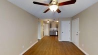This single-floor space presents an open, connected layout that seamlessly links the main living area to the kitchen. Warm hardwood flooring extends throughout, adding a sense of continuity and warmth to the interior. The living area is equipped with a ceiling fan featuring a built-in light fixture, providing both air circulation and ambient lighting. Neutral-toned walls complement the flooring and contribute to a clean, understated aesthetic.

Toward the rear, the kitchen is visible and features gray cabinetry paired with a classic white subway tile backsplash, creating a timeless and practical design. A stainless steel appliance is integrated beneath the countertop, and the sink is positioned along the back wall beneath the cabinetry. A small chandelier-style light fixture illuminates the kitchen area. A hallway corridor connects the living space to additional areas of the home, suggesting further rooms beyond what is immediately visible. Two white paneled doors are also present, likely leading to a closet and an additional room or exterior exit. White baseboards frame the space cleanly throughout.