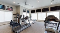This fitness center offers a comprehensive and well-appointed workout environment designed to support a variety of exercise routines. The space is outfitted with a selection of cardio machines, including treadmills and an elliptical, alongside strength training equipment such as a weight bench and resistance machines. A dedicated storage rack keeps accessories and free weights neatly organized and within reach.

Natural light fills the room through multiple large windows dressed with white shades, creating a bright and inviting atmosphere. A ceiling fan overhead helps maintain a comfortable temperature during workouts. Multiple large flat-screen televisions are mounted along the upper walls, allowing members to stay entertained or informed while exercising.

The flooring is a durable, low-pile carpet suited for fitness use, and the neutral, light-toned walls contribute to an open and clean aesthetic. Recessed lighting supplements the natural light, ensuring the space remains well-lit at any time of day. Motivational framed artwork adorns the walls, adding a polished finishing touch. Overall, this fitness center combines functionality with a welcoming design, providing everything needed for a productive and enjoyable workout.