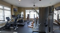 This fitness center offers a comprehensive workout environment designed to accommodate a wide range of exercise needs. The space is thoughtfully laid out across a single floor, making efficient use of the available area. Large windows line one wall, flooding the room with natural light and providing an open, airy atmosphere. A ceiling fan overhead ensures comfortable air circulation during workouts.

The facility is outfitted with an impressive selection of equipment. Cardio machines, including a treadmill and stationary bike by Cybex, are positioned along the mirrored wall, which spans a generous portion of the room and visually expands the space. Strength training options include a multi-function cable machine and a dedicated leg press or extension station with tan upholstered padding, along with additional resistance equipment.

Durable dark rubber flooring covers the ground, providing safety and stability for all types of exercise. Recessed lighting supplements the natural light, ensuring the space remains well-lit at all times. A wall-mounted flat-screen television adds an entertainment option for those seeking distraction during cardio sessions. The neutral wall tones and clean layout give the gym a professional yet welcoming ambiance suited for regular fitness use.