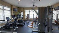 This fitness center offers a comprehensive workout environment designed to accommodate a wide range of exercise needs. The space is thoughtfully laid out across a single floor, making efficient use of the available area. Large windows line one wall, flooding the room with natural light and providing an open, airy atmosphere. A ceiling fan overhead ensures comfortable air circulation during workouts.

The facility is outfitted with an impressive selection of equipment. Cardio machines, including a treadmill and stationary bike by Cybex, are positioned along the mirrored wall, which spans a generous portion of the room and visually expands the space. Strength training options include a multi-function cable machine and a dedicated leg press or extension station with tan upholstered padding, along with additional resistance equipment.

Durable dark rubber flooring covers the ground, providing safety and stability for all types of exercise. Recessed lighting supplements the natural light, ensuring the space remains well-lit at all times. A wall-mounted flat-screen television adds an entertainment option for those seeking distraction during cardio sessions. The neutral wall tones and clean layout give the gym a professional yet welcoming ambiance suited for regular fitness use.