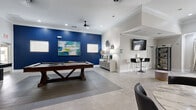 Located at 50 Estuary Trail in Alpharetta, Georgia, this single-floor community space is thoughtfully designed to offer both recreation and relaxation. The interior is finished with light-toned tile flooring throughout, crown molding, and recessed lighting, lending the space a clean, modern aesthetic.

The centerpiece of the space is a generously sized game room featuring a full-size billiards table set against a bold navy accent wall adorned with contemporary artwork. A ceiling fan overhead ensures comfort during play, while natural light filters in through multiple windows. Adjacent to the billiards area, a shuffleboard table and a bistro-height table with bar seating provide additional entertainment options, complemented by a wall-mounted flat-screen TV.

The lounge area offers a spacious, open layout with a large sectional sofa, decorative ottomans, and curated wall art and mirrors that add depth and elegance to the room. A bay window area with floor-length curtains provides a bright, welcoming atmosphere. A nearby reading nook features a cushioned accent chair, a shelving unit stocked with books and décor, and a sunny window—ideal for quiet moments.

Together, these areas create a well-rounded community gathering space that balances social entertainment with comfortable, inviting design.