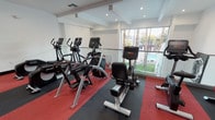 This fitness center is a well-appointed, single-floor facility designed to support a comprehensive cardio workout experience. The space features a clean, modern aesthetic with crisp white walls, recessed lighting, and a coffered ceiling detail that adds architectural interest overhead. Durable red and black rubberized flooring lines the workout area, providing both safety and a bold visual contrast to the light surroundings.

The facility is equipped with a robust selection of Life Fitness cardio machines, including multiple treadmills, elliptical cross-trainers, and upright and recumbent stationary bikes. The equipment is thoughtfully arranged to maximize the use of space while maintaining an open, uncluttered feel.

Natural light floods the interior through expansive floor-to-ceiling glass windows that line two sides of the room, offering views of a sparkling outdoor pool area surrounded by mature palm trees and lush greenery. A mirrored wall further enhances the sense of openness, reflecting both light and the surrounding outdoor scenery. The combination of generous natural light, garden views, and well-maintained professional equipment creates an inviting and motivating environment well-suited for daily exercise.