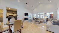 This single-floor clubhouse is designed to serve as an inviting and functional hub for community gatherings. The centerpiece of the space is a generously sized main lounge area defined by dramatic vaulted ceilings that lend an airy, open feel throughout. Gleaming large-format tile flooring runs seamlessly across the main areas, complementing the bright white walls and recessed lighting that keep the space feeling clean and welcoming.

The main lounge is anchored by a classic fireplace with a white mantle, topped by a wall-mounted television — creating a cozy yet social focal point. Multiple seating arrangements, including plush neutral sofas and accent chairs, are thoughtfully arranged to encourage conversation and relaxation. French doors with transom windows line one wall, allowing an abundance of natural light and providing views to the exterior.

A casual dining area with a glass-top table and upholstered chairs sits adjacent to the lounge, ideal for community meals or informal meetings. Nearby, a kitchenette features upper cabinetry, a counter-height bar with seating, and essential amenities for hosting events.

A separate, more intimate room with rich hardwood flooring, a formal dining table, and large windows offering natural light provides a versatile private meeting or dining space. Together, these areas make the clubhouse a well-rounded and comfortable community amenity.