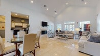 This single-floor clubhouse is designed to serve as an inviting and functional hub for community gatherings. The centerpiece of the space is a generously sized main lounge area defined by dramatic vaulted ceilings that lend an airy, open feel throughout. Gleaming large-format tile flooring runs seamlessly across the main areas, complementing the bright white walls and recessed lighting that keep the space feeling clean and welcoming.

The main lounge is anchored by a classic fireplace with a white mantle, topped by a wall-mounted television — creating a cozy yet social focal point. Multiple seating arrangements, including plush neutral sofas and accent chairs, are thoughtfully arranged to encourage conversation and relaxation. French doors with transom windows line one wall, allowing an abundance of natural light and providing views to the exterior.

A casual dining area with a glass-top table and upholstered chairs sits adjacent to the lounge, ideal for community meals or informal meetings. Nearby, a kitchenette features upper cabinetry, a counter-height bar with seating, and essential amenities for hosting events.

A separate, more intimate room with rich hardwood flooring, a formal dining table, and large windows offering natural light provides a versatile private meeting or dining space. Together, these areas make the clubhouse a well-rounded and comfortable community amenity.