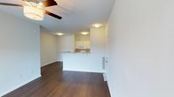 This single-floor residence offers an open and airy layout that seamlessly connects the living area to the kitchen. The space is finished with rich, dark hardwood flooring throughout, providing a warm contrast to the crisp white walls and ceilings. Flush-mount ceiling lights illuminate the interior evenly, while a ceiling fan in the living area adds both style and comfort.

The kitchen is positioned centrally within the floor plan and features a peninsula with a light-toned countertop that doubles as a breakfast bar, creating a natural division between the kitchen and living spaces without sacrificing the open feel. White cabinetry lines the kitchen walls, and appliances including a microwave are integrated cleanly into the design. A sink is visible along the counter, and the layout allows for efficient workflow.

The living area is generously proportioned, offering flexibility in furniture arrangement. Clean baseboards and neutral wall tones throughout create a move-in-ready aesthetic. A door visible along the wall suggests access to additional rooms or a utility area. The overall ambiance is bright, clean, and modern, making this a practical and welcoming space well-suited for everyday living.