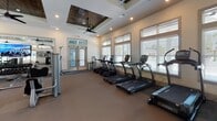Located at 50 Cheryl Ln in Summerville, SC, this fitness center is a thoughtfully designed, single-floor facility that combines elegant architectural details with a comprehensive selection of exercise equipment. The space is defined by its striking coffered tray ceilings with rich wood plank inlays, recessed lighting, and multiple ceiling fans that maintain airflow and comfort throughout. Large windows with plantation-style blinds line the walls, flooding the interior with abundant natural light and offering views of the surrounding greenery. The main workout floor is generously sized and features a wide variety of strength-training machines, including cable systems, functional trainers, benches, and a power rack, providing options for a full-body workout. A dedicated row of treadmills is positioned along the windowed wall, making for an enjoyable cardio experience. Additional cardiovascular equipment includes stationary bikes, a stair climber, and a rowing machine. Large wall-mounted flat-screen televisions are positioned throughout the space. A separate room with glass doors houses a dedicated spin studio with multiple cycling bikes. Floor-to-ceiling mirrors expand the sense of space, while framed black-and-white athletic photography adds a motivating aesthetic touch. The overall ambiance is bright, open, and well-appointed.