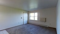 This single-floor space presents a straightforward and functional layout centered around a generously sized main room. The room features wall-to-wall carpeting in a neutral gray tone, providing a practical and comfortable underfoot surface. Smooth, light-colored walls create a clean backdrop throughout the space, complemented by a plain ceiling that maintains a simple, unadorned aesthetic.

Natural light enters the room through a standard double-pane window, offering a view of the surrounding outdoor environment. A six-panel white interior door with brown trim provides access to an adjoining area or room. The space is equipped with baseboard heating running along the lower perimeter of the wall, as well as a wall-mounted radiator unit, indicating a dual heating setup suited for colder climates.

An electrical outlet is visible along the baseboard, and a small wall-mounted device — possibly a thermostat or intercom — is positioned near the door. The overall ambiance is minimal and neutral, making the space highly adaptable for a variety of uses. The combination of practical flooring, functional heating systems, and adequate natural light contributes to a livable and versatile environment.