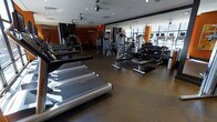 This fitness center offers a comprehensive workout environment designed to accommodate a wide range of exercise needs. The space is laid out across a single floor, with a spacious open floor plan that allows for easy movement between equipment zones. A row of treadmills lines one side of the room, providing dedicated space for cardio training. Adjacent to the cardio area, a well-stocked free weights section features a multi-tiered dumbbell rack paired with adjustable benches, ideal for strength training routines. A selection of cable and resistance machines occupies the rear of the space, offering additional options for targeted muscle training. The flooring throughout is a durable rubber surface, providing both safety and comfort during workouts. Full-length mirrors span one wall, enhancing the sense of space while allowing users to monitor their form. Large floor-to-ceiling windows along the exterior wall flood the gym with natural light and offer views of the surrounding streetscape. The ceiling is fitted with recessed lighting and ceiling fans, ensuring consistent illumination and airflow. Bold orange accent walls add a dynamic, energizing visual contrast to the otherwise neutral palette, contributing to the motivating atmosphere of the facility.