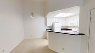 This single-floor residence offers an open and airy layout that seamlessly connects the living area to the kitchen. The living space is distinguished by notably high ceilings that amplify the sense of volume, complemented by a ceiling fan that aids in air circulation throughout the room. Soft, neutral-toned carpeting covers the living area floor, contributing to a warm and understated ambiance.

The kitchen is designed around a wraparound peninsula with a dark contrasting countertop, providing both workspace and a natural visual divider between the cooking and living areas. White cabinetry lines the kitchen walls, offering generous storage, while a skylight positioned above floods the workspace with natural light. White appliances, including a refrigerator and range, maintain a cohesive, clean aesthetic throughout the kitchen. The overall color palette of the space is consistently light and neutral, with white walls and trim reinforcing a bright, open feel. The single-floor layout ensures easy accessibility and a straightforward flow throughout the residence.