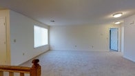 This single-floor space presents a generously sized open room finished with wall-to-wall carpet in a neutral grey-beige tone, providing a clean and versatile foundation. The room is defined by smooth, white walls and a flat ceiling, giving it a simple, understated aesthetic throughout. A window fitted with horizontal blinds allows natural light to filter into the space, brightening the interior while offering privacy control. The ceiling features a centrally located HVAC vent, indicating a ducted climate control system for year-round comfort. Multiple electrical outlets are distributed along the walls, offering practical utility for various furniture arrangements and uses. A wooden staircase newel post is partially visible in the foreground, suggesting access to a lower or upper level nearby. The overall ambiance of the space is clean, minimal, and move-in ready, offering a blank canvas suitable for a range of configurations. The neutral palette and open layout make it highly adaptable, whether intended for use as a living area, recreation room, or additional flex space.