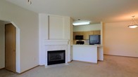 This single-floor residence offers a well-designed open-concept layout that seamlessly connects the living and kitchen areas. The living room is anchored by a classic white-framed fireplace, which serves as a striking focal point and adds warmth and character to the space. Soft, neutral carpeting throughout the living area contributes to a comfortable and inviting ambiance.

The kitchen is positioned adjacent to the living room and is accessible via a raised breakfast bar that doubles as a pass-through, creating a sense of connectivity while maintaining a distinct cooking zone. The kitchen is equipped with light oak cabinetry, black appliances including a refrigerator, range, and microwave, and features overhead fluorescent lighting that keeps the workspace well-lit. The breakfast bar provides a casual dining or prep surface, enhancing the kitchen's functionality.

The neutral color palette of warm whites and beiges used throughout the space creates a clean, cohesive aesthetic that feels both open and easy to personalize. The layout is efficient and practical, making thoughtful use of the available space. Overall, the design balances everyday functionality with a welcoming residential atmosphere suited for comfortable living.