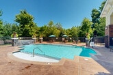 Located at Potomac Heights Place in Woodbridge, Virginia, this beautifully designed outdoor amenity area centers around a generously sized swimming pool with crystal-clear blue water. The pool deck is finished in warm stamped concrete, providing an inviting and durable surface for lounging and socializing. Stone-accented pillar features and decorative planters with lush tropical foliage add elegant architectural detail throughout the space.

The pool is complemented by a charming clubhouse structure that blends red brick and natural stone masonry with cream-colored siding and a standing-seam metal roof. Large windows on the clubhouse facade allow natural light to flow freely, while a covered portico entry provides a welcoming transition between indoor and outdoor spaces.

The surrounding deck area is generously appointed with outdoor seating, including table-and-chair sets shaded by teal market umbrellas, ideal for relaxation or casual gatherings. A decorative stone water feature wall serves as a focal point at one end of the pool area, enhancing the resort-like ambiance.

The entire outdoor space is enclosed by a black metal fence, ensuring privacy and security. Mature trees lining the perimeter provide natural shade and a lush green backdrop, contributing to the serene, park-like atmosphere of this well-maintained community amenity in Woodbridge, VA.